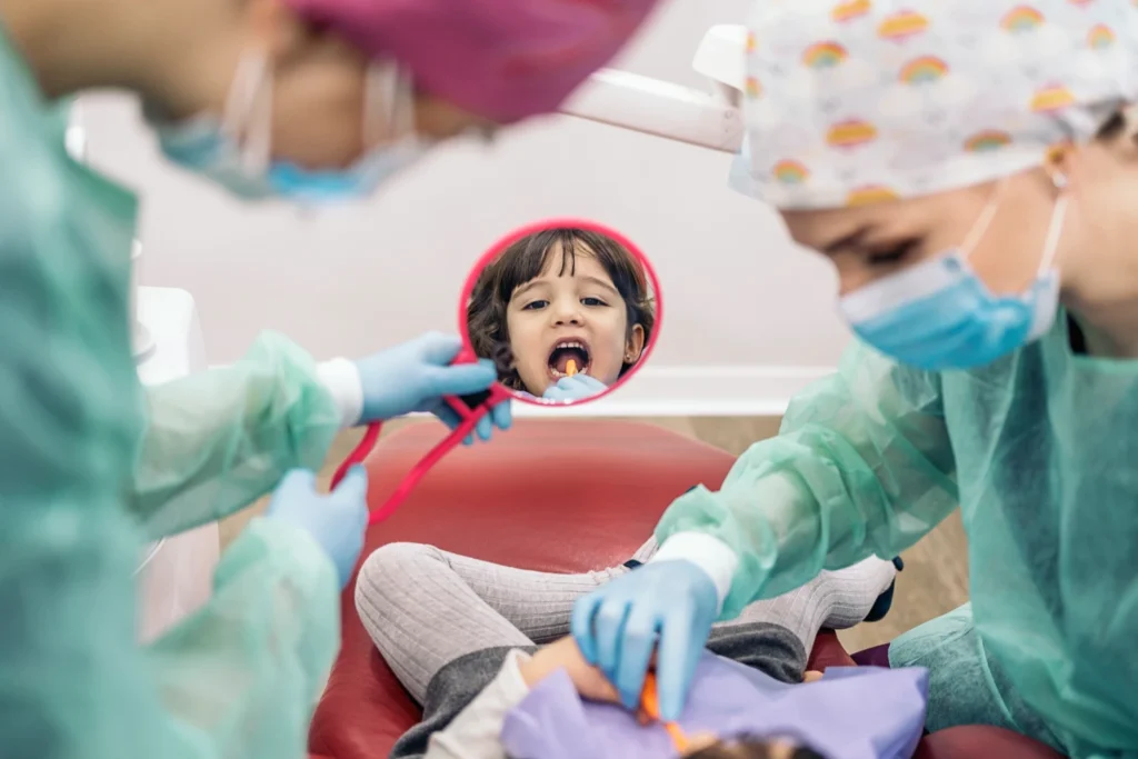 Niña en consulta dental, mostrando su boca abierta mientras dos dentistas la examinan. Uno de los dentistas sostiene un espejo que refleja la cara de la niña. Ambos profesionales llevan guantes y mascarillas, y están vestidos con batas de protección. La escena transmite un ambiente amigable y profesional en la atención odontológica infantil.