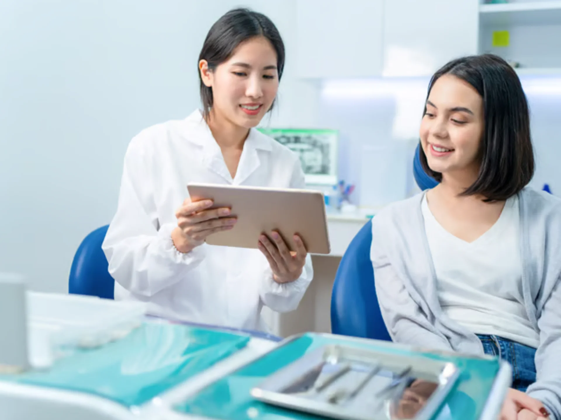 Dentista explicando tratamiento a paciente en clínica dental, con tablet en mano y una mesa de instrumentos dentales al fondo.