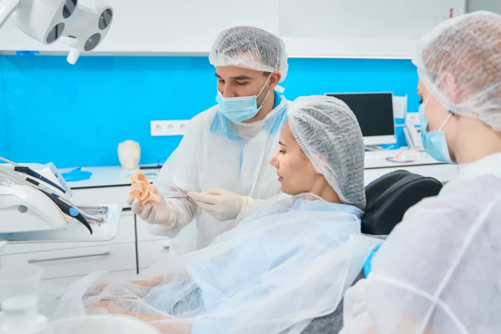 Dentista mostrando un modelo dental a una paciente en un consultorio odontológico moderno, con personal usando equipo de protección y un ambiente limpio y profesional.